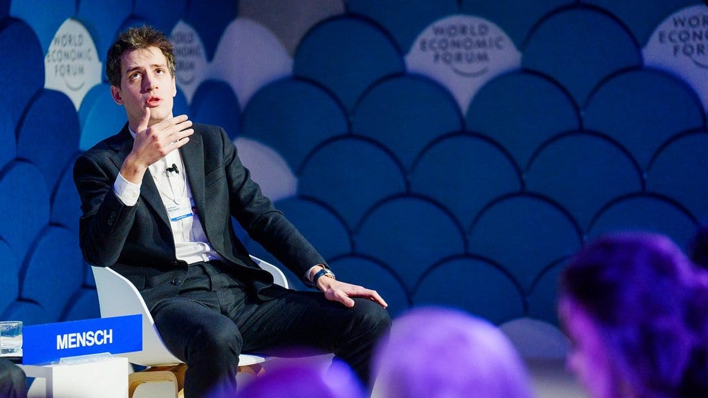Arthur Mensch sits on a panel stage in front of a blue backdrop, a nameplate reading Mensch beside him.