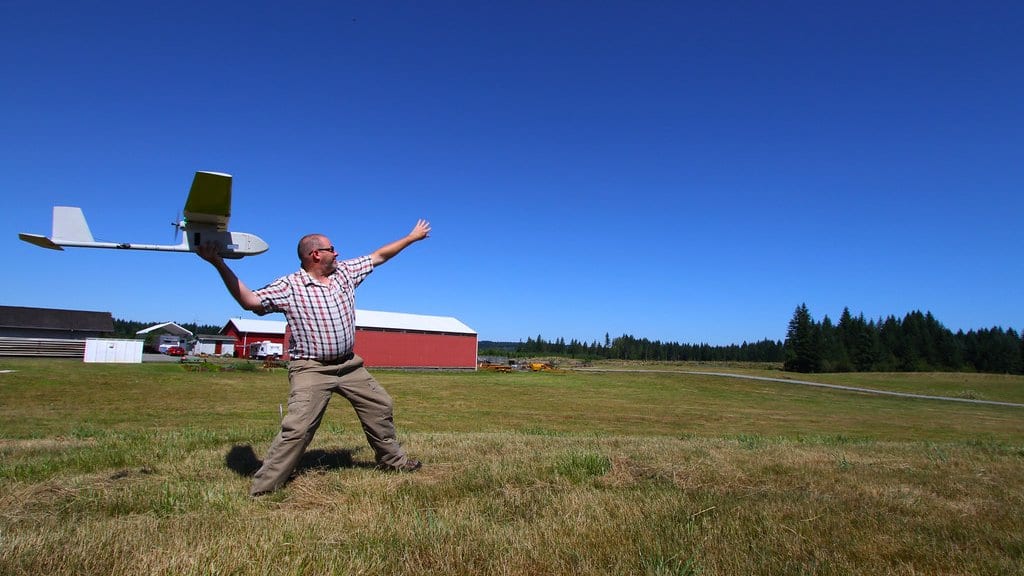 A researcher hand launches a small white fixed-wing uncrewed aircraft over a grass field under a clear blue sky.