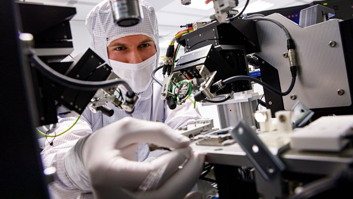 A researcher in a white clean suit and mask works at an optical laboratory bench, reaching between microscope objectives, micropositioners and cables around a small optical component.