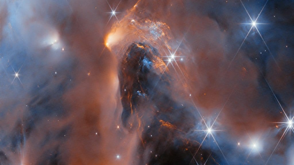 Infrared image of glowing orange and blue dust clouds, a dark pillar of dust and bright spiked stars in a star-forming cloud.