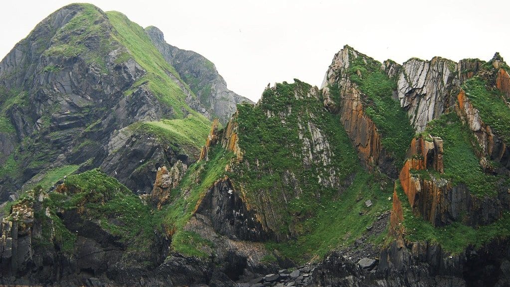 Steep green and orange sea cliffs of sharply tilted rock layers rising straight out of the ocean in the Shumagin Islands, Alaska.