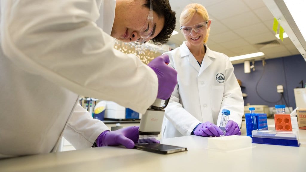Two researchers in white coats at a laboratory bench, one lowering a multichannel pipette onto a multiwell plate.