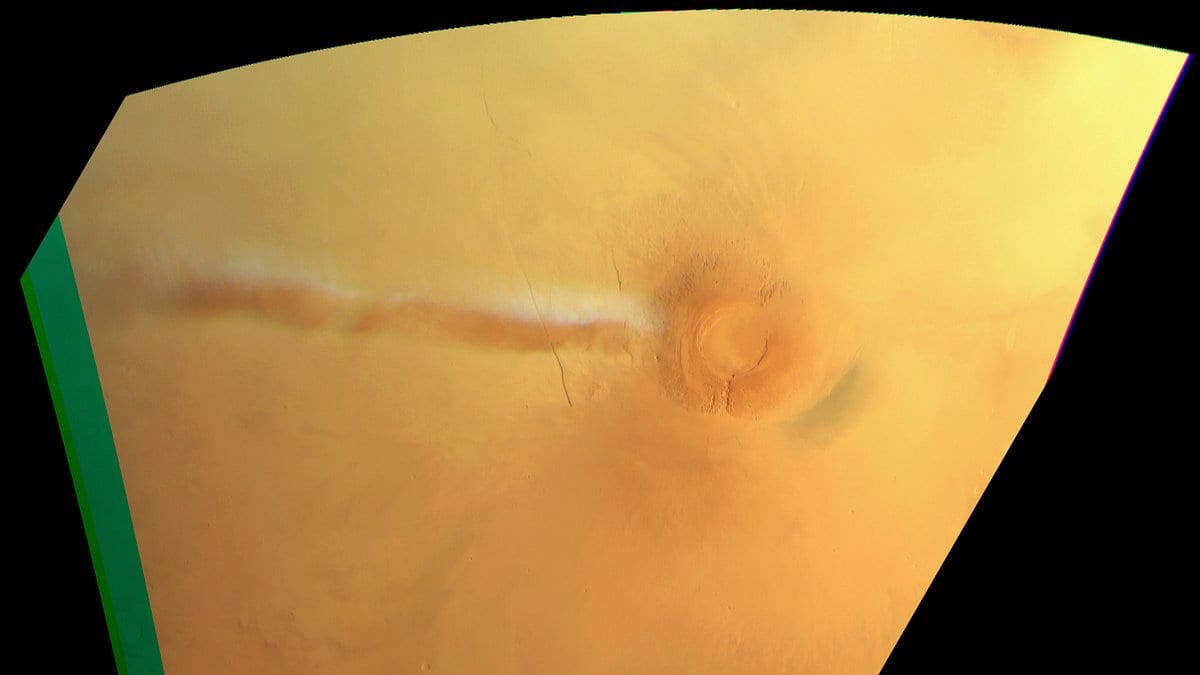 A long white water-ice cloud stretches west across the orange Martian surface from the dark summit caldera of the Arsia Mons volcano.