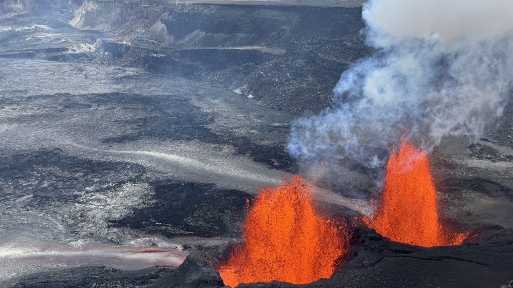 Two orange lava fountains erupt from a vent on the dark floor of a large volcanic crater, with a steam plume rising and the caldera wall behind.