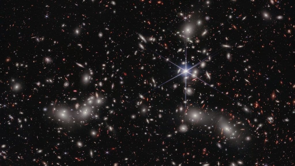 A dense field of hundreds of galaxies in many shapes and colors on a black sky, with one bright foreground star showing eight-pointed diffraction spikes.
