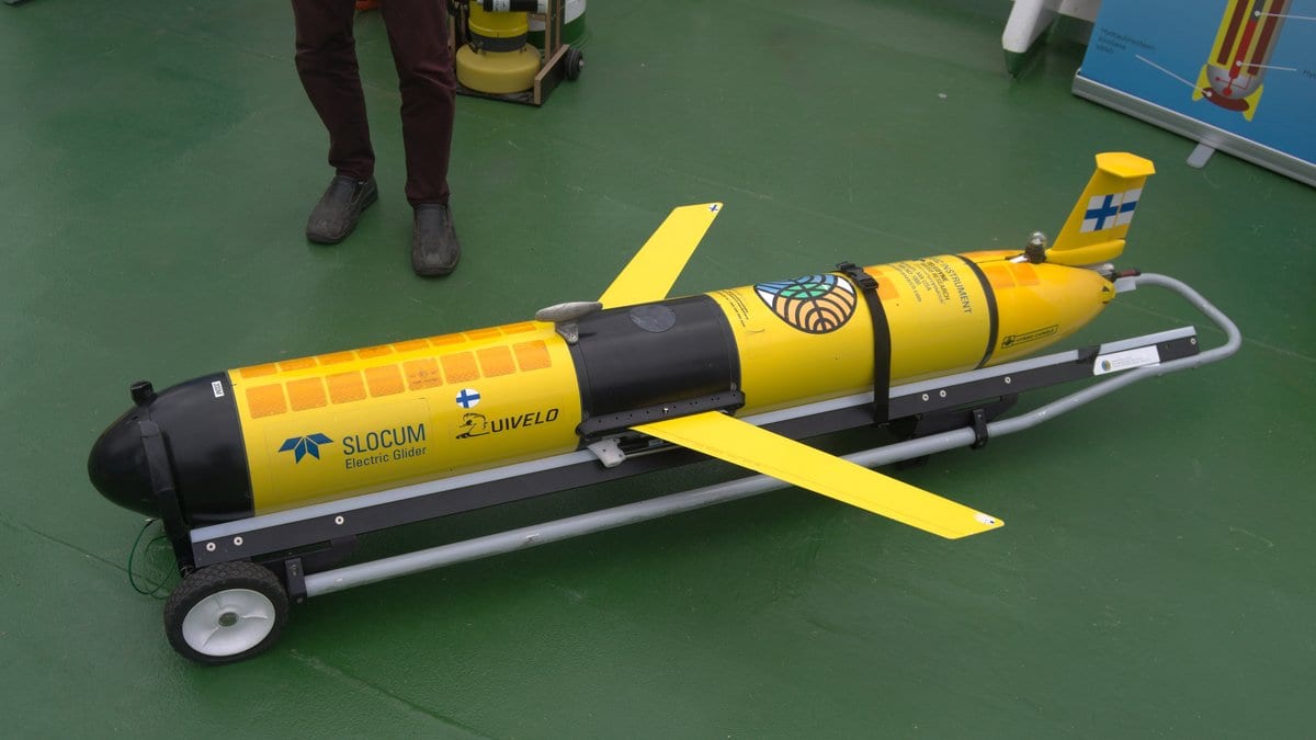 A yellow and black autonomous underwater glider with swept wings and a tail fin, resting on a wheeled cradle on a green deck.