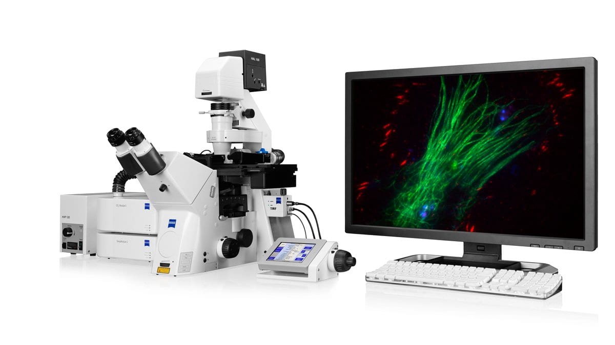 A commercial inverted fluorescence microscope fitted with a total internal reflection module, beside a monitor showing a multicolor fluorescence image of cells.