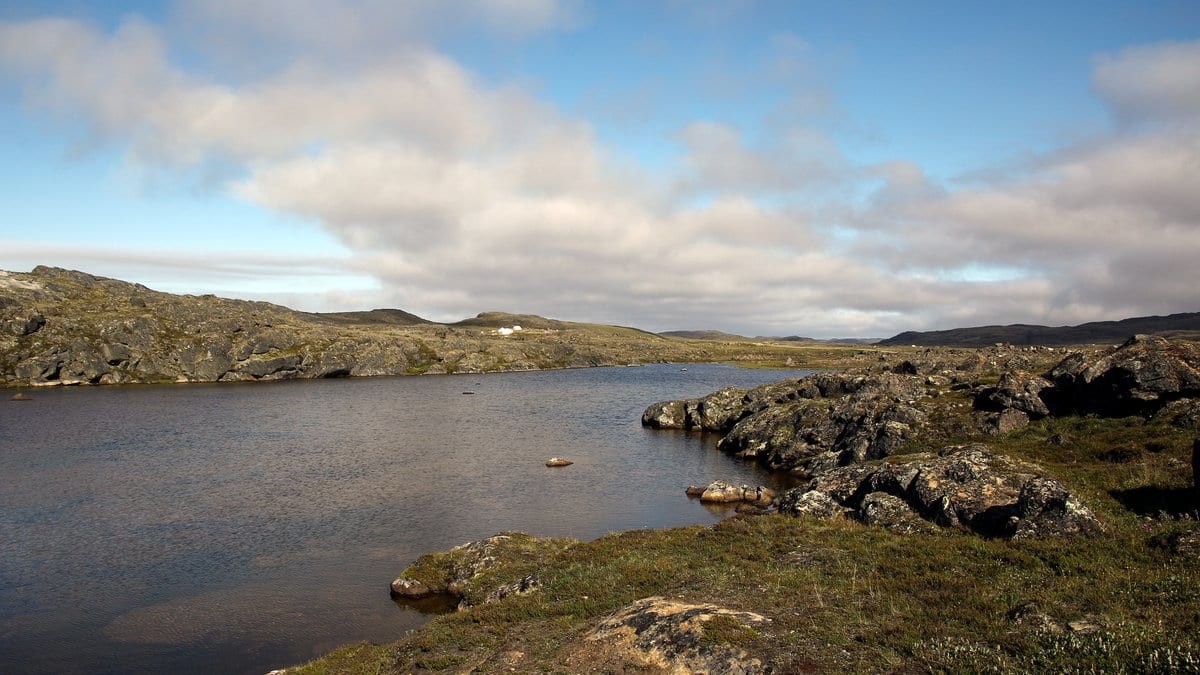Calm fresh water among rock outcrops and low tundra under a partly cloudy sky on Baffin Island, Nunavut.
