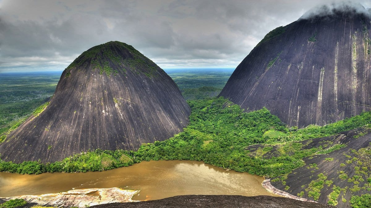 A wide brown river winds between two rounded granite domes covered in rainforest under a heavy gray sky.