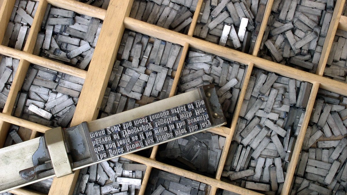 Compartments of a wooden printing case filled with loose metal type, with an assembled line of type lying across them.