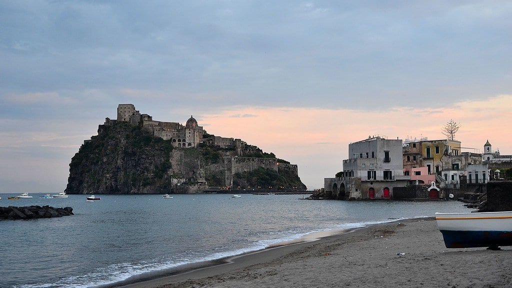 A fortified rocky islet rising from the sea off Ischia in the Gulf of Naples, seen from a nearby beach at dusk.