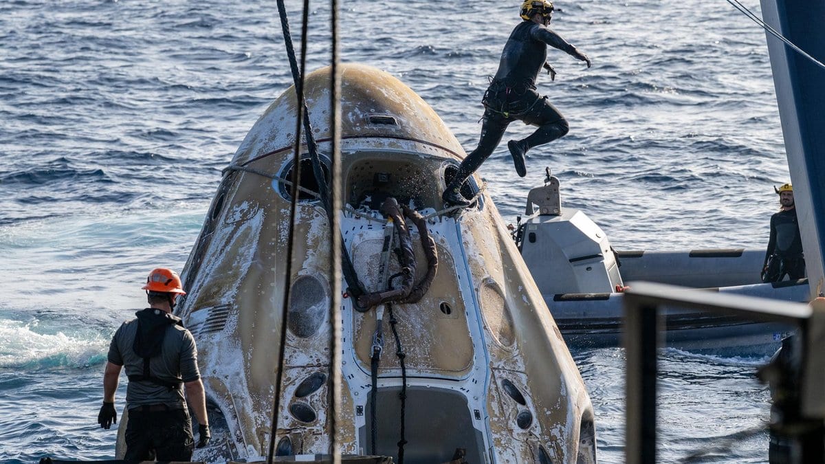 A SpaceX Crew Dragon capsule floats in the ocean next to a recovery boat while two crew members work on the hull, one jumping down onto the spacecraft.