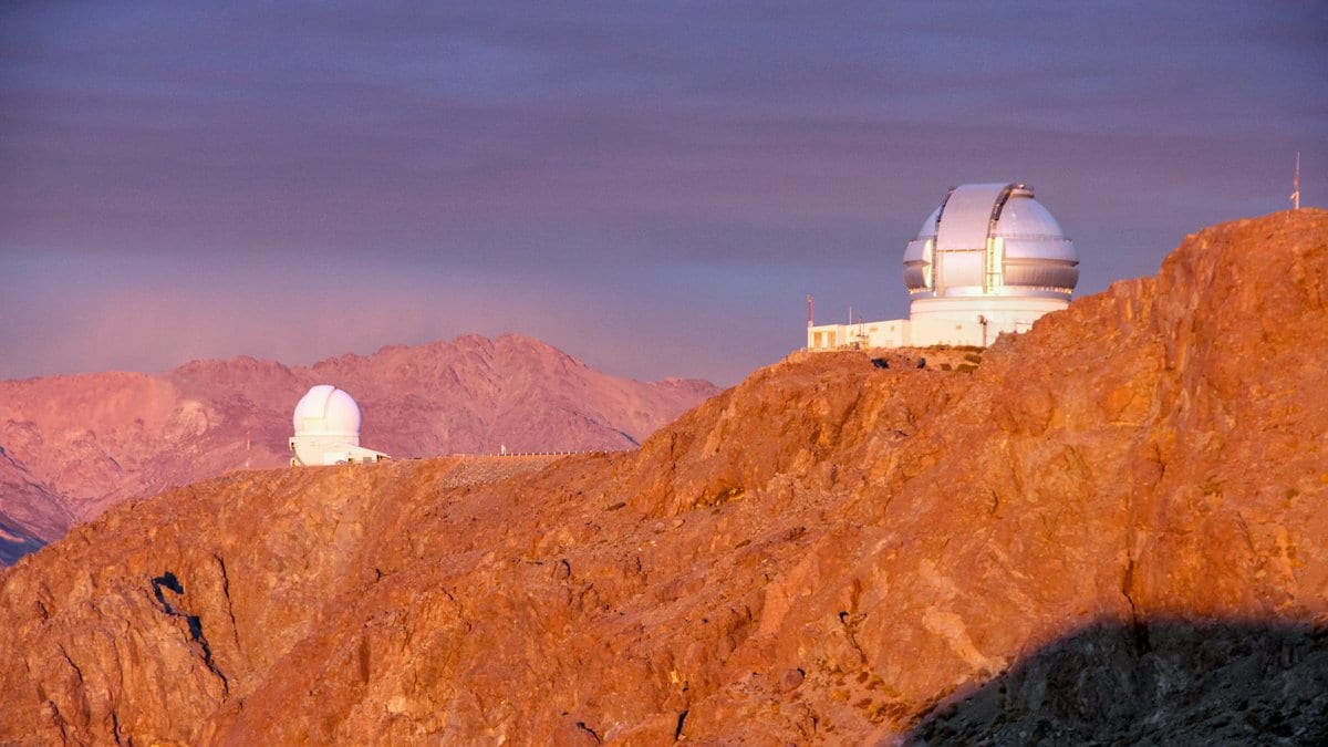 Two white observatory domes on a rocky Andean ridge, lit orange by low sunlight, with hazy mountains behind.