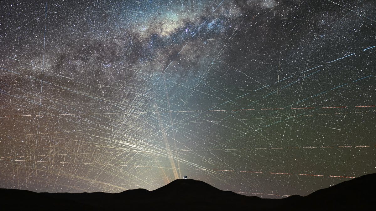 Dozens of straight satellite trails criss-cross a star-filled night sky above a dark desert ridge, with the Milky Way overhead and a small observatory dome silhouetted on the horizon.