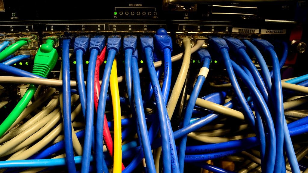 Blue, beige, green, yellow and red patch cables plugged into the front ports of a rack mounted ethernet switch, with link lights glowing between them.