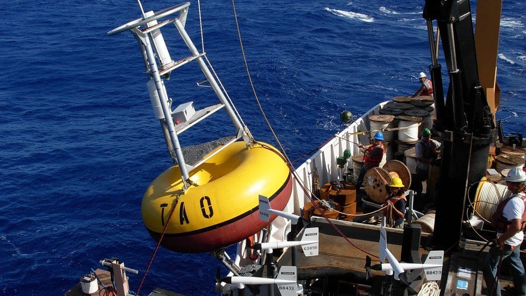 A yellow and red TAO moored buoy hangs from a ship crane above deep blue tropical ocean while crew in hard hats work on the deck below.