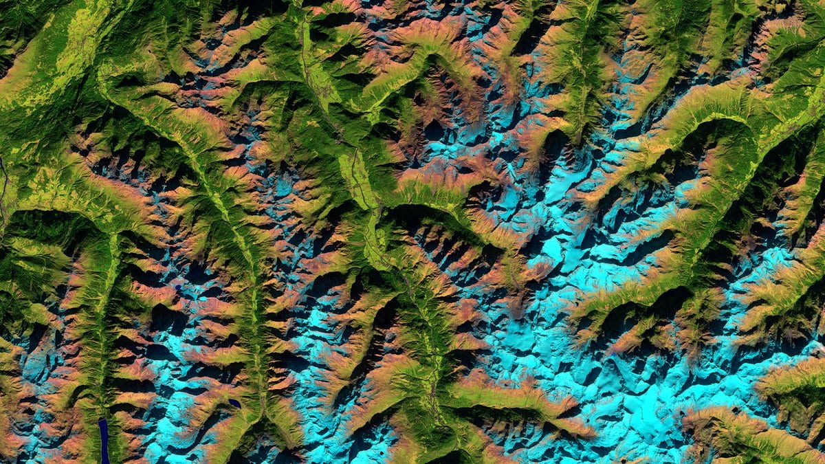 False-color satellite view of a steep mountain range, with narrow ridges and deep valleys in green and brown and the highest ground in pale cyan.
