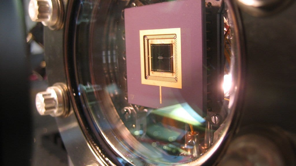A microfabricated ion-trap chip mounted behind the glass viewport of a vacuum chamber, surrounded by bolted metal flanges.