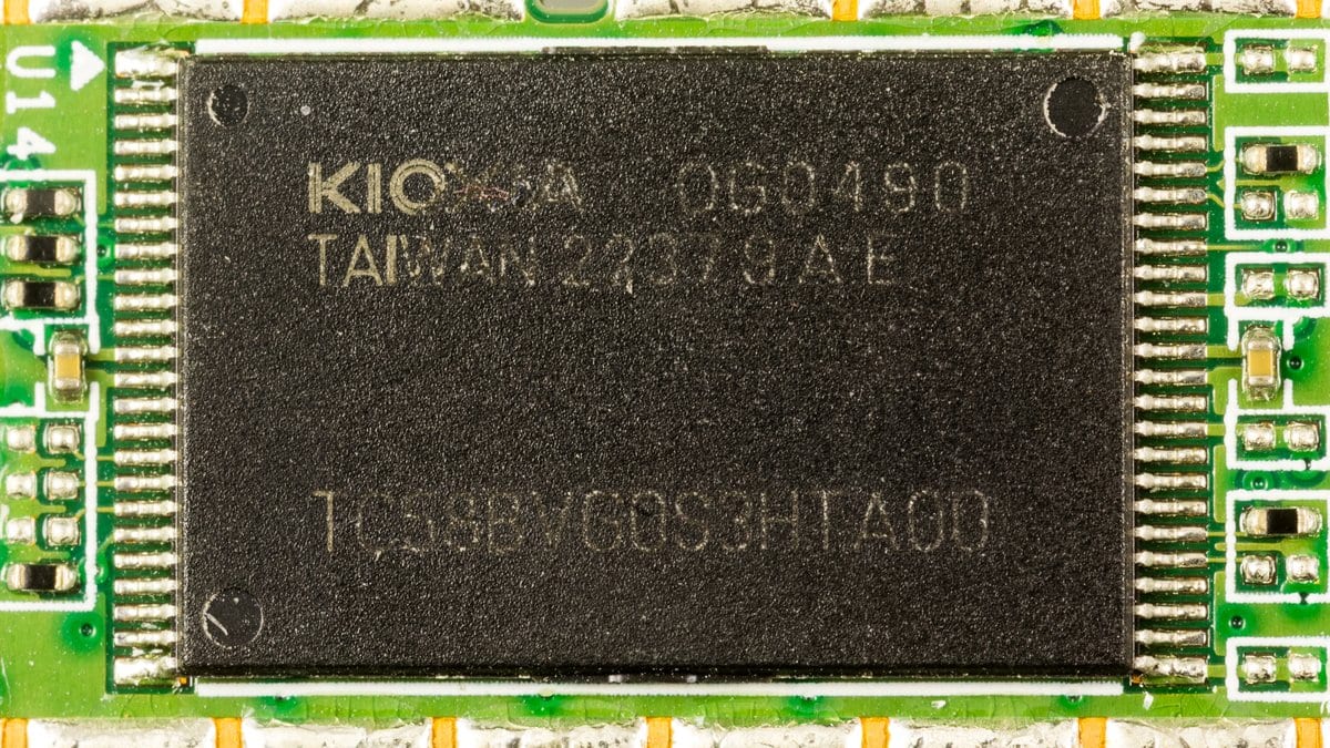 A black Kioxia NAND flash memory package soldered to a green circuit board, with its part number printed on the chip.
