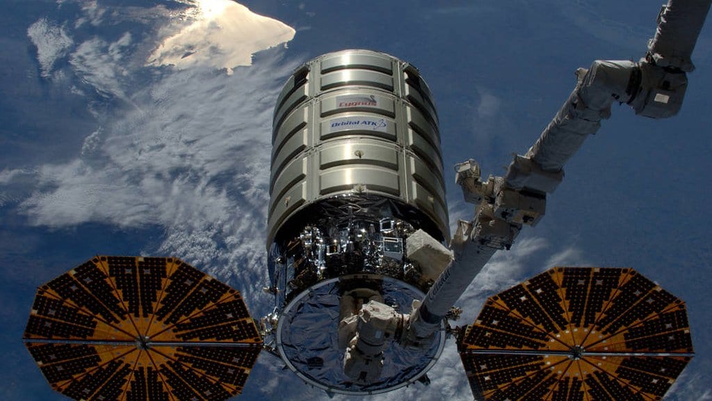 A barrel-shaped Cygnus cargo spacecraft with two round gold solar arrays, held by the International Space Station's robotic arm above a cloud-covered ocean.