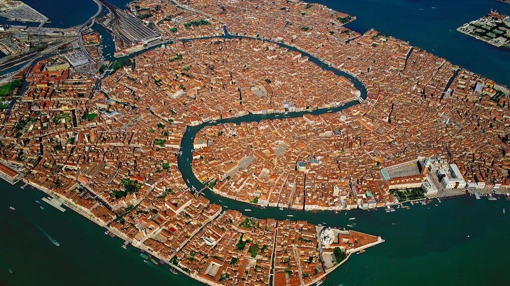 Aerial view of central Venice on its islands, ringed by the shallow water of the lagoon, with the Grand Canal curving through the city.