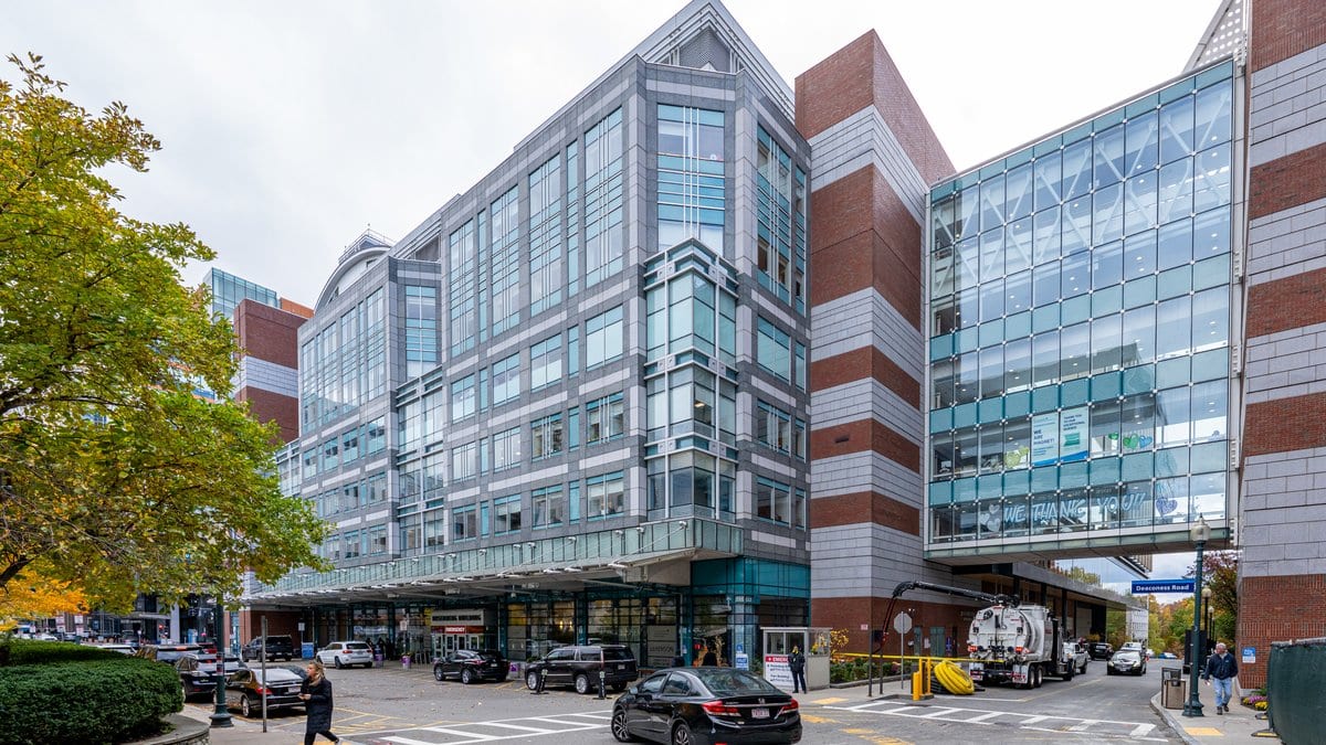 Street view of the glass and brick buildings of Beth Israel Deaconess Medical Center in Boston, with cars and a walkway at the main entrance.
