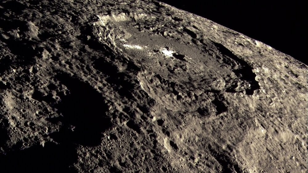 Oblique view from orbit of a large crater on the gray, heavily cratered surface of the dwarf planet Ceres, with a bright white deposit on its floor and the curve of the limb against black space.