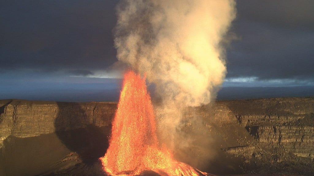 A tall orange lava fountain erupting from a vent on the floor of Kīlauea's summit crater, throwing a gas and ash plume above the layered crater walls.