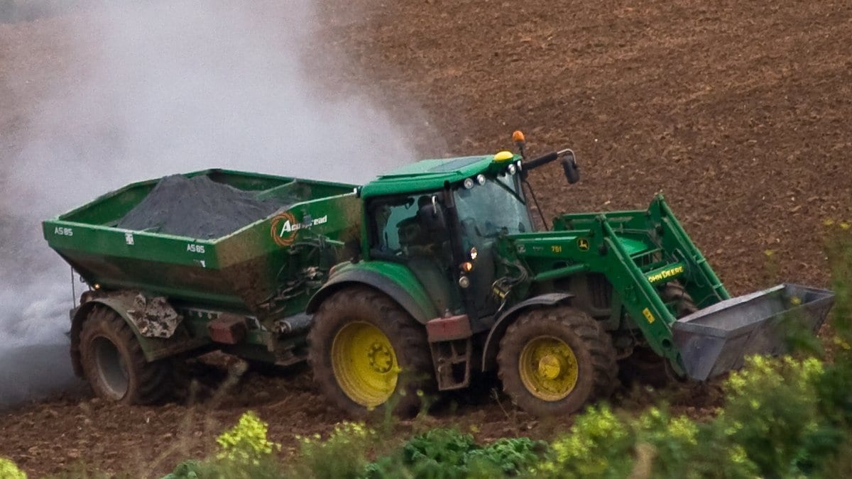 A green tractor pulls a hopper spreader across a plowed field, trailing a pale plume of dust.