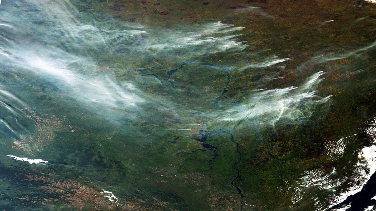 Satellite view of gray wildfire smoke drifting over dark green Siberian forest, with river channels visible through the haze.