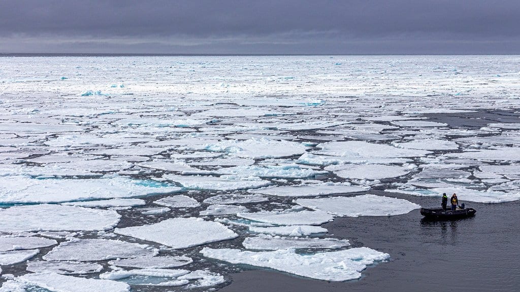 Flat white sea ice floes scattered across dark Arctic Ocean water under a gray sky, with a small inflatable boat carrying two people at the right.