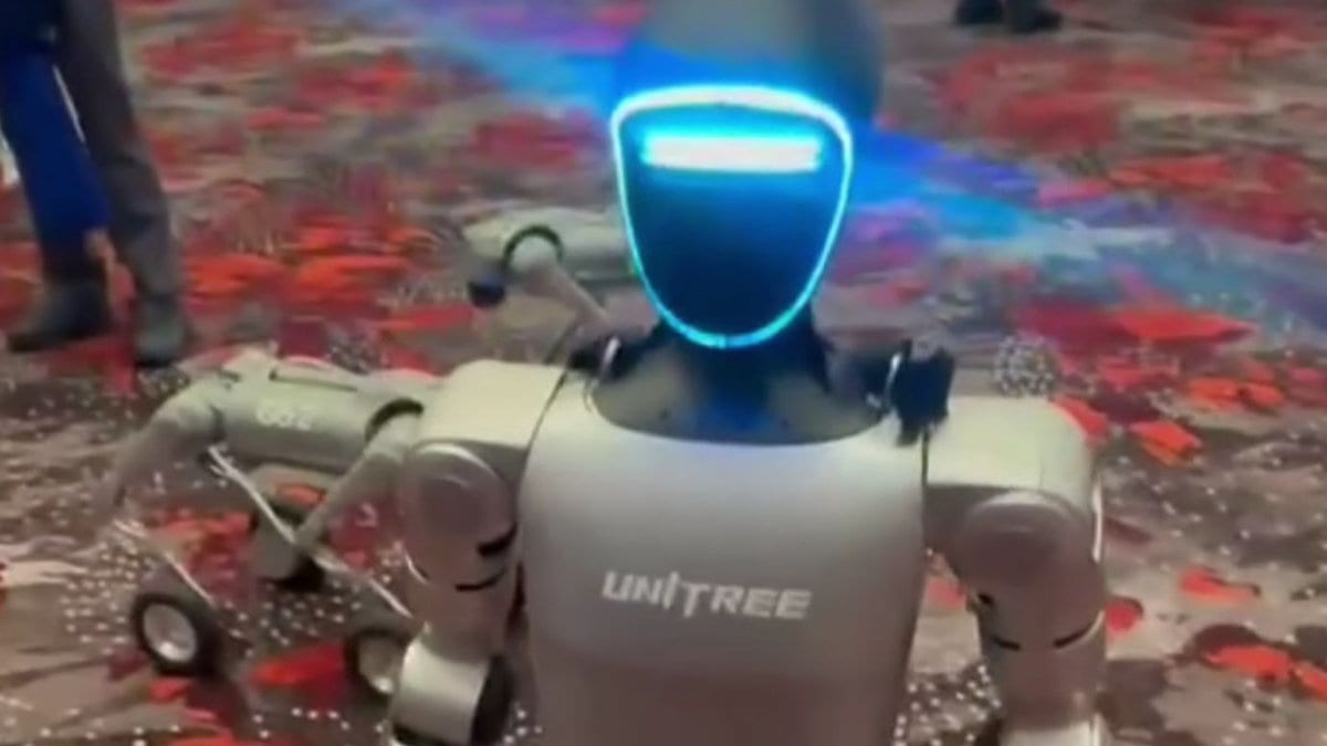 A silver humanoid robot with a glowing blue visor and UNITREE lettering across its chest, walking across a patterned carpet with a four-legged robot behind it.