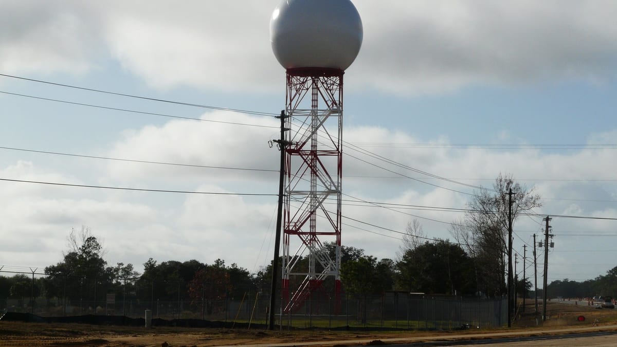 A large white radar dome on a red and white lattice tower behind a chain-link fence, power lines and an empty road in the foreground