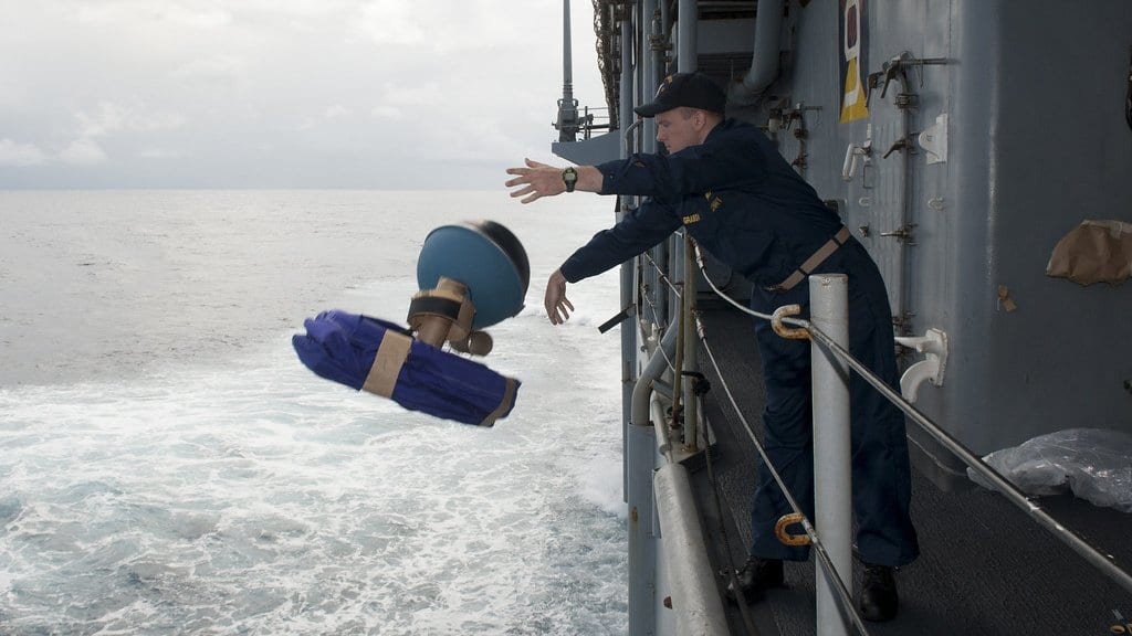 A sailor leans over a ship's rail and releases a drifting buoy, its folded drogue trailing below the float, toward the sea.