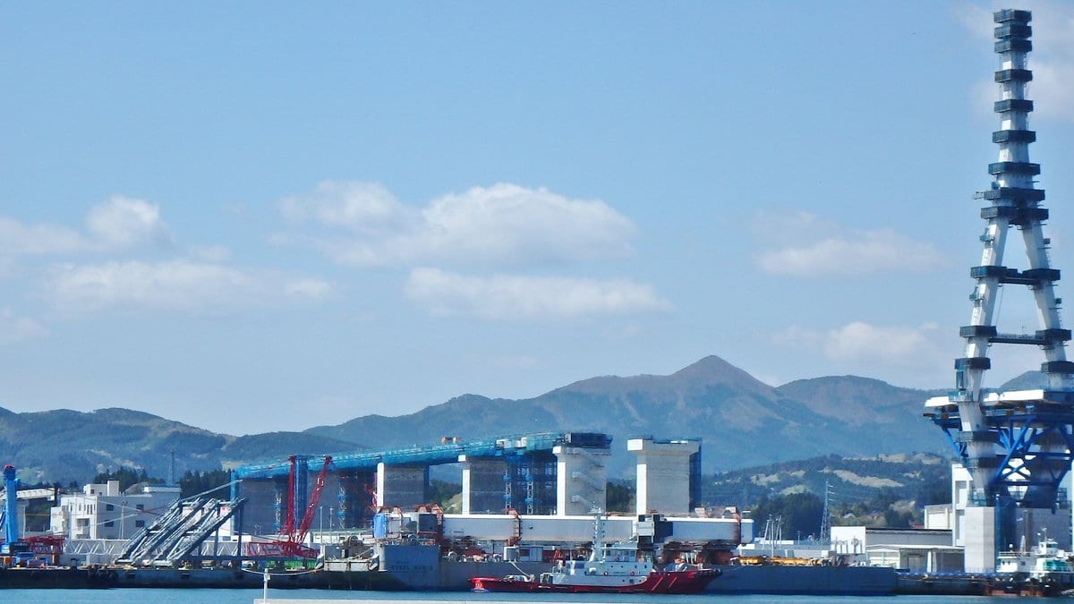 Daytime view across Kesennuma Bay toward the working port, with cargo wharves, red construction cranes, a tall steel bridge tower on the right and forested mountains behind.