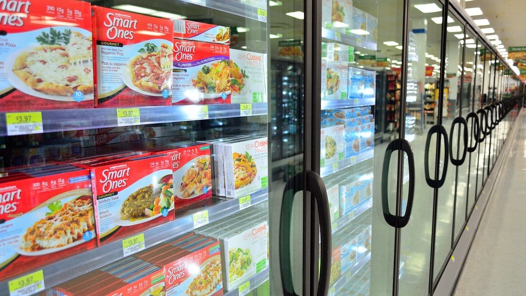 A long row of glass-door freezer cabinets stocked with packaged frozen meals, receding down a supermarket aisle.