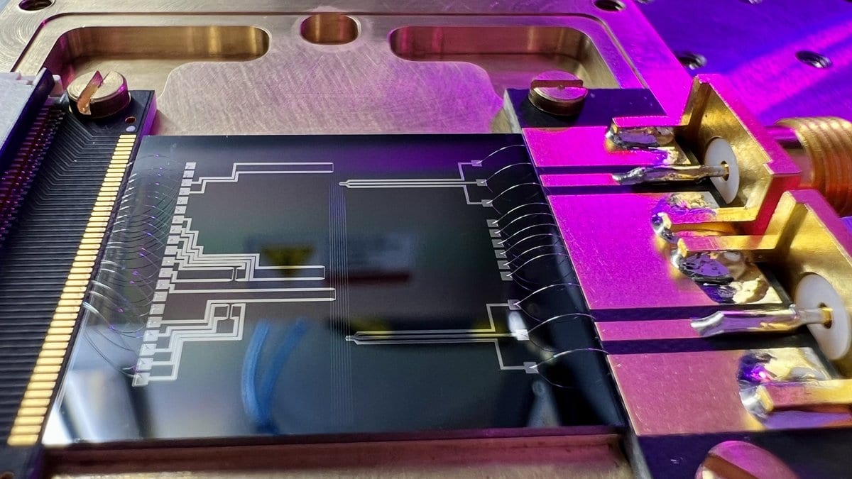 Close view of an integrated photonic chip wire bonded to a gold mount, its waveguide circuit visible as fine pale lines across the dark surface under purple laboratory lighting.