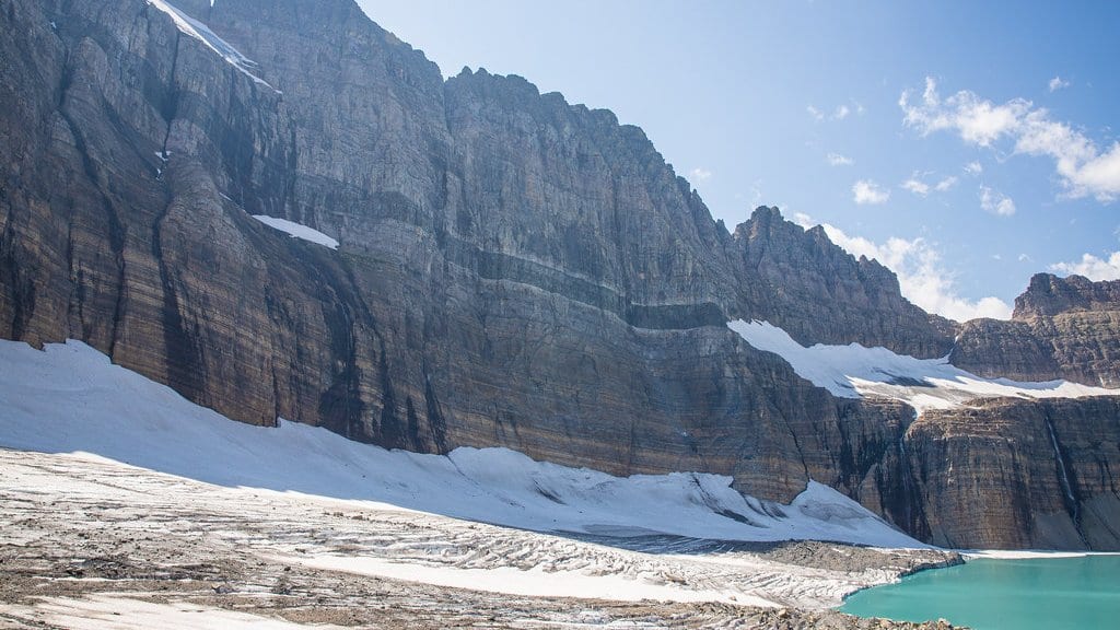 A broad field of snow and glacier ice at the base of a tall layered rock cliff, with a turquoise meltwater lake at lower right, in Glacier National Park, Montana.