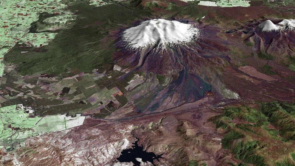 Satellite view of Mount Ruapehu in New Zealand, a snow-covered summit cone ringed by dark bare volcanic slopes and braided valleys, with green farmland to the west.