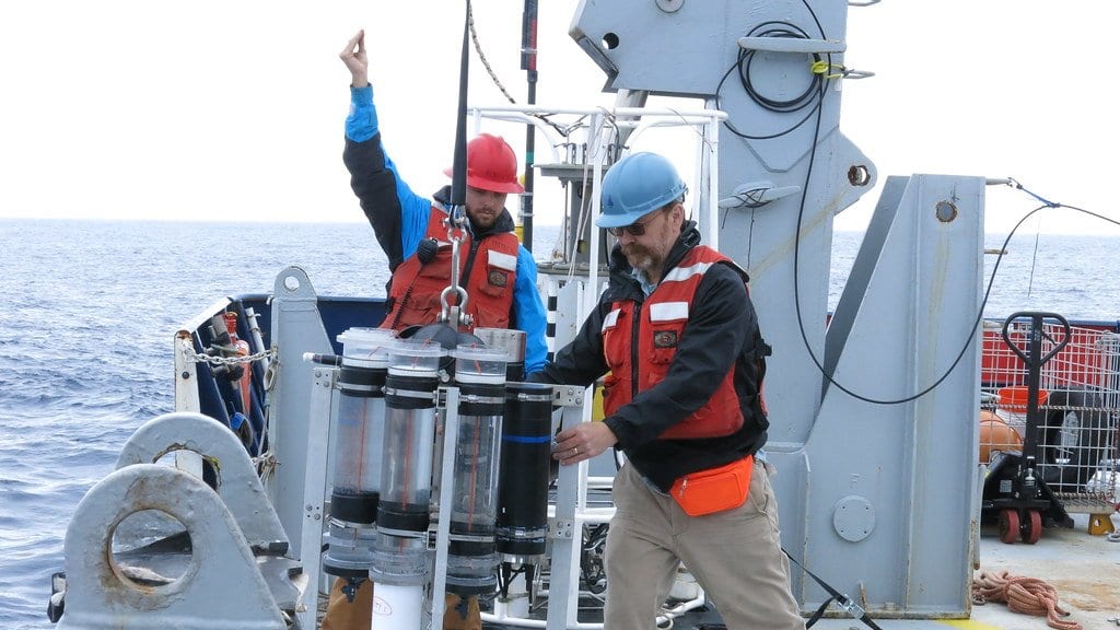 Two researchers in hard hats and life vests lower a cylindrical sediment trap over the side of a research ship at sea.