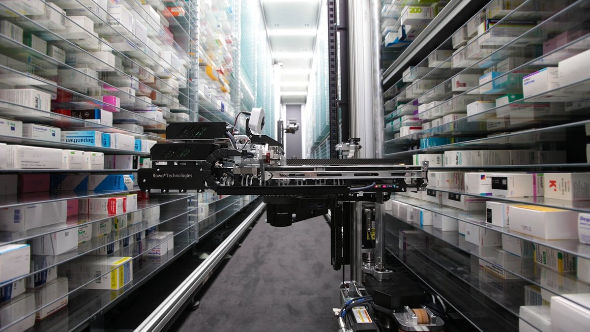 An automated pharmacy dispensing robot travels on a rail down a narrow aisle between tall racks of boxed medicines.