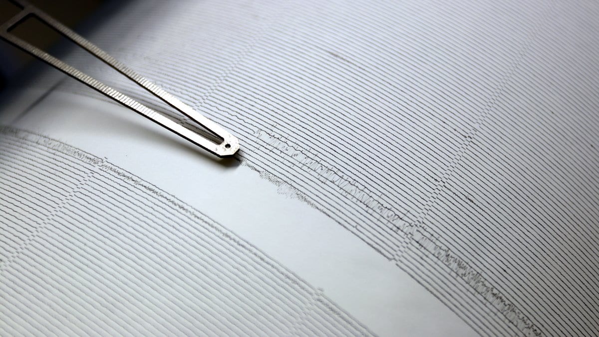 Close-up of a seismograph stylus arm resting on a paper drum record covered in closely spaced wavy ink lines.