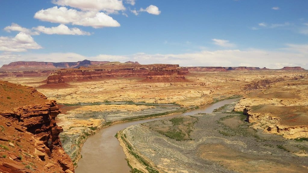 A muddy river curves through a wide desert canyon of red and tan rock under a blue sky with scattered clouds.