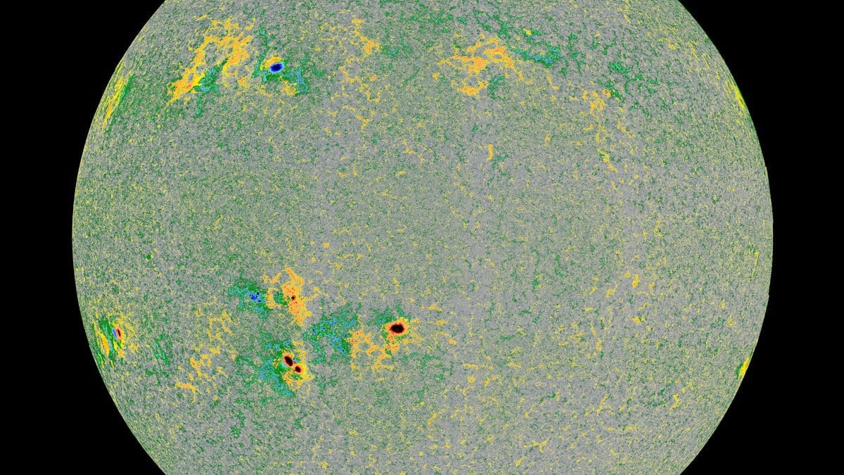 A full-disk magnetic map of the Sun, mottled gray with yellow and green patches where the field is strongest, on a black background.
