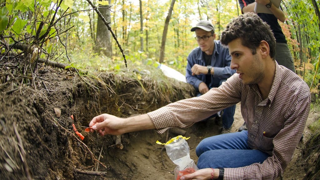 Two researchers kneel beside an open pit in a forest, placing small colored flags into the exposed soil face.