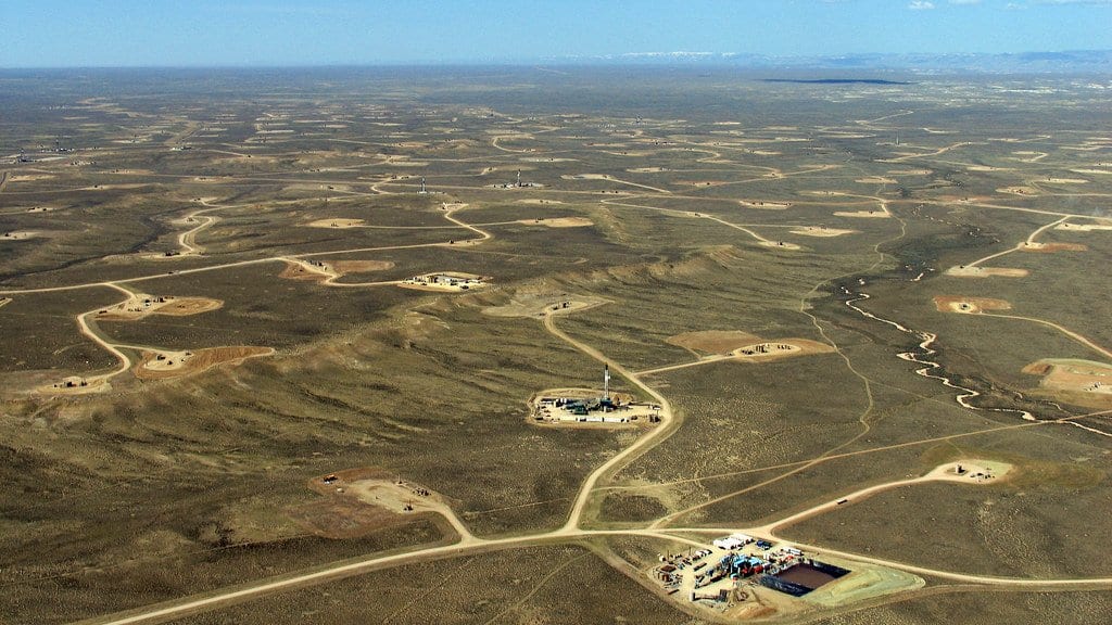 Aerial view of a natural gas field in western Wyoming, with dozens of cleared well pads linked by dirt access roads across sagebrush plains.