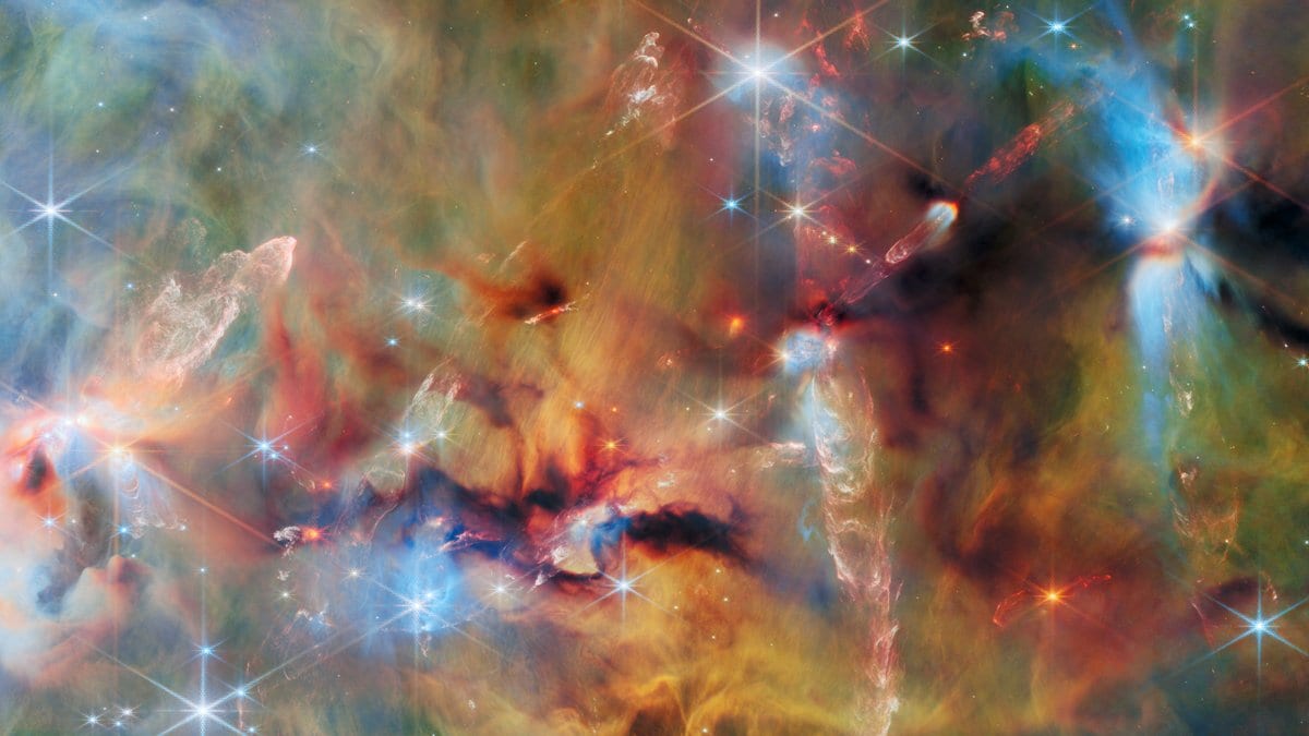 A wide near-infrared view of a star-forming cloud, with glowing orange, green and blue filaments of gas and dust, dark dust lanes, and many bright stars showing eight-pointed diffraction spikes.