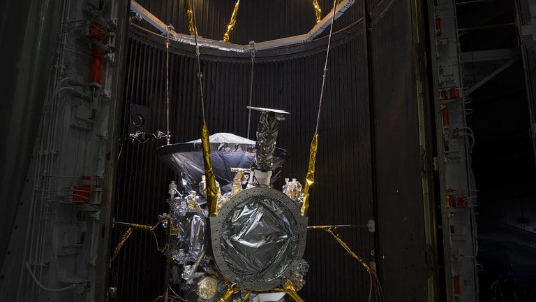 A spacecraft covered in gold and silver thermal blankets stands inside a tall cylindrical vacuum chamber whose heavy door is swung open.