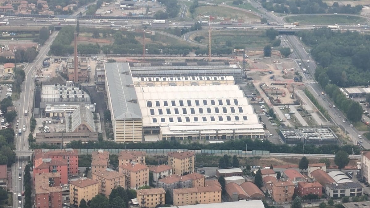 An aerial photograph of the European Centre for Medium-Range Weather Forecasts site in Bologna, Italy, a long hall with a white roof beside rail lines and apartment blocks.