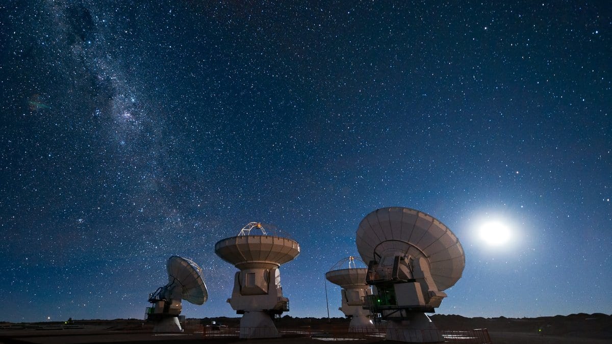 Four ALMA radio antennas on the Chajnantor plateau in Chile under a dense starry sky, with the Moon low on the right.